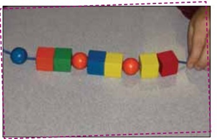 A sequence of wooden beads makes a necklace. From left to right: blue sphere, orange cube, green cube, orange sphere, blues cube, yellow cube, orange sphere, yellow cube and red cube.