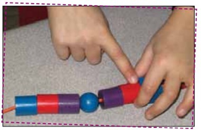 A sequence of wooden beads makes a necklace. From left to right: blue cylinder, red cylinder, purple cylinder, blue sphere, purple cylinder, red cylinder, and blue cube.