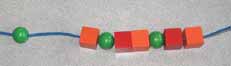 A sequence of wooden beads makes a necklace. 3 elements ae repeated 3 times, green sphere, red cube, orange cube. At the beginning of the necklace a red cube is missing where there is a spacing. 