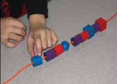 The hands of a student show the first element of a repetitive motif: blue cylinder, purple cylinder, red cylinder, repeated 3 times.