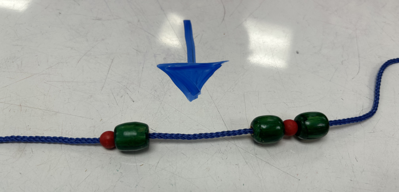 A sequence of repetitive patterns repeated 3 times, beads forming a necklace. The pattern is red oval bead, a green sphere bead. A space is left to show where the missing bead should go. A blue arrow is used to show the space.