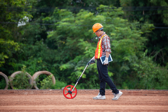 Un homme portant un casque et un gilet orange marche avec un odomètre à roue. 