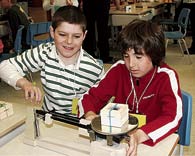 Two children measure the weight of an object using a triple beam scale.