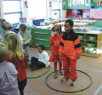 Classroom situation. There are two hoops on the floor. In the first hoop, one child is standing and the other is sitting. In the second hoop, both children are standing.  There are other children in the classroom.