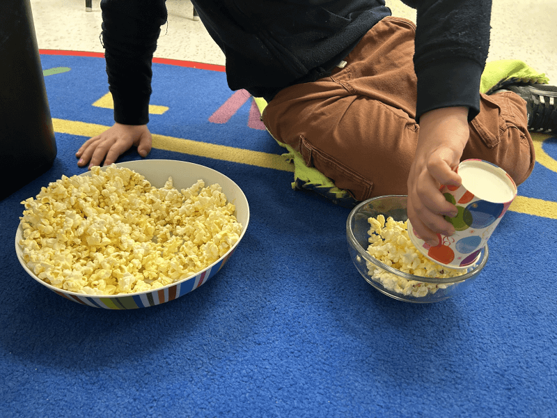 A student uses a paper cup to transfer popcorn from one container to another