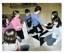 Students are sitting on the floor, manipulating large cardboard figures.