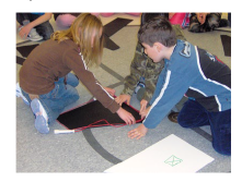 Students are sitting on the floor, manipulating large cardboard shapes.